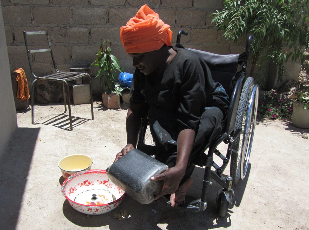 A woman seated in a wheelchair wearing a black top and orange head covering is pouring water into a bowl.