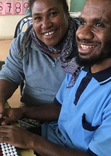 This photo shows a Papua New Guinean woman and man sitting next to each other, using an abacus and smiling. The women is an Inclusive Education teacher from an Inclusive Education Resource Centre and is teaching a student who is blind how to make mathematical calculations.