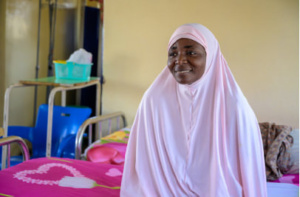 A Nigerian woman wearing a pale pink khimar sitting on a hospital bed.
