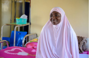 A Nigerian woman wearing a pale pink khimar sitting on a hospital bed.