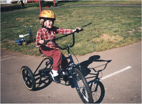 A young boy wearing red trousers and a red checkered shirt and a yellow helmet while riding a tricycle.