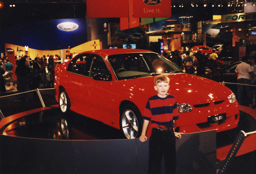 A young boy wearing a black and red striped top standing in front of a red Holden car.