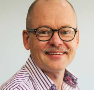 Rev Dr Kirk Patston wearing a striped shirt and black framed glasses.