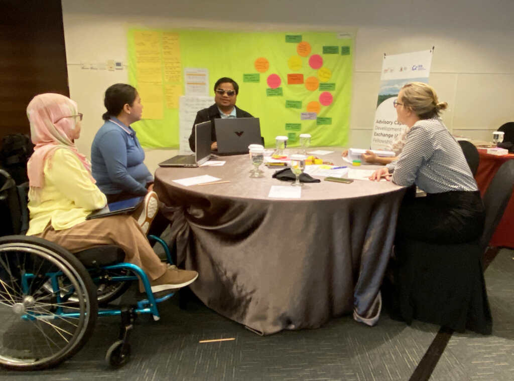 Four persons sitting around a round table with a tablecloth in front of a board with post its. One woman on the left is sitting in a wheelchair and is wearing a headscarf. The woman next to her has dark hair in a ponytail and is wearing a blue shirt. The man at the back of the table is wearing envision glasses, has dark hair and wears a dark jacket. The woman on the right front has blond hair, wears glasses and a light shirt.