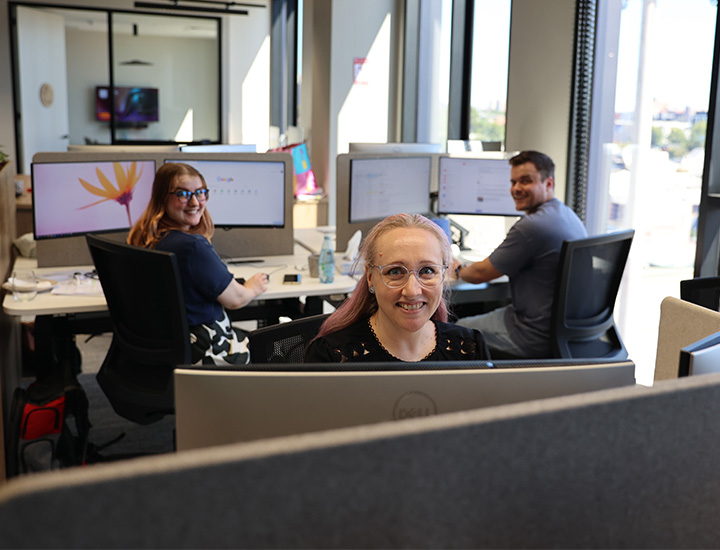 Two women and a man seated at computers smiling towards the camera. One woman has glasses and a purple dress, the other has glasses and is in a black dress. The man is wearing a gray shirt and jeans.