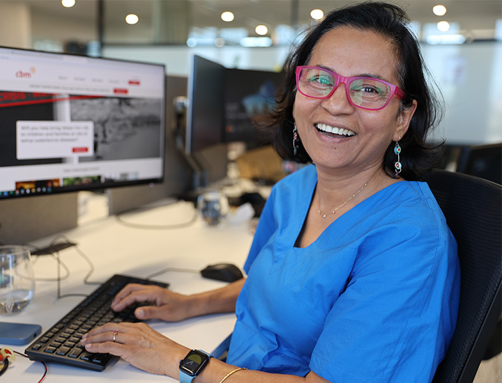 A woman with black hair and glasses seated at a computer. She is wearing a blue dress.