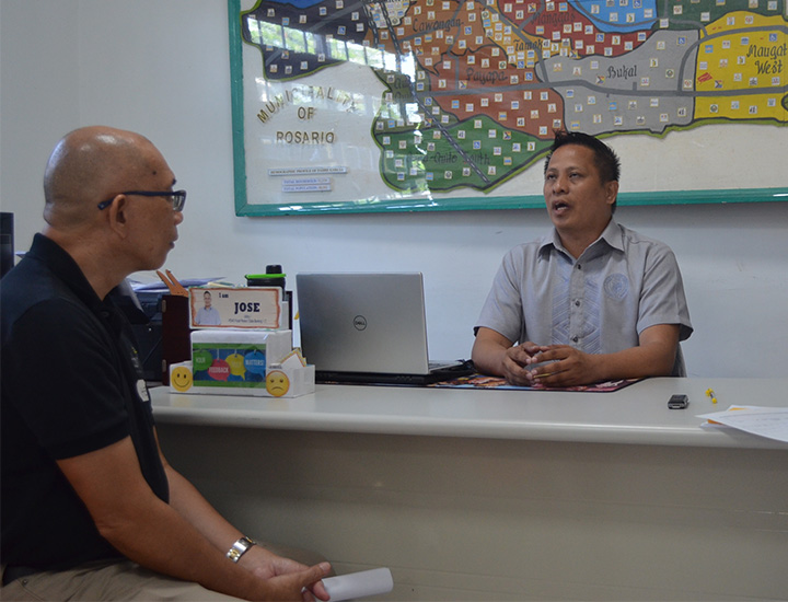 A man in a button-up shirt sits at a desk, speaking to a man in a black polo shirt and glasses, in an office with a 'Municipality of Rosario' sign and map on the wall.