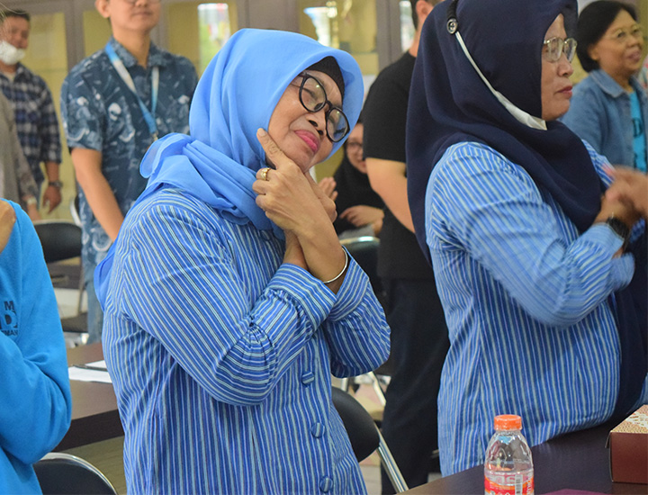 Two women in blue striped uniforms and head coverings making the sign language sign for smiling.