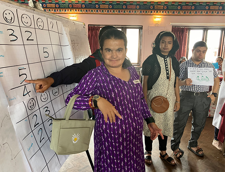 Two women and a man standing in a room. One woman has a purple flowery dress, the other has a white dress and black cardigan. The man with Down syndrome is holding a piece of paper that reads 'I love easy-to-read material group discussions' in Nepali.