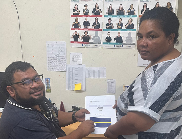 In an office with a sign language guide on the wall, a man in glasses and a polo shirt, and a woman in a white and grey shirt, together hold up a 'Visit Program' document from the Consulate General of Sydney, Australia.