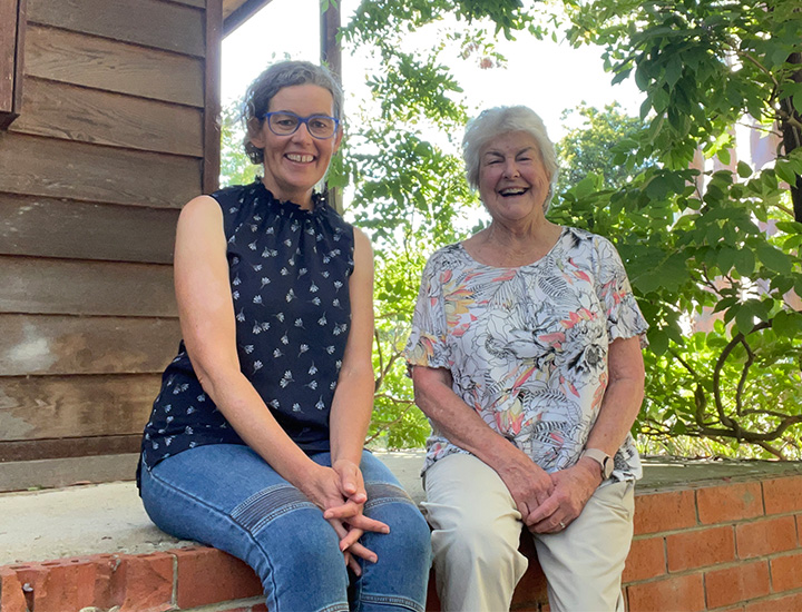 Two woman sitting beside one another smiling towards the camera. The woman on the left has glasses and is in a purple shirt and blue jeans. The woman on the right is in a white shirt and beige pants.