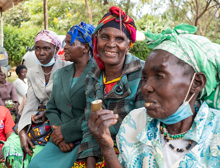 Four Kenyan women in headwraps, two of them with face masks, one of them having one eye, standing outdoors among other people. One of them smiles.