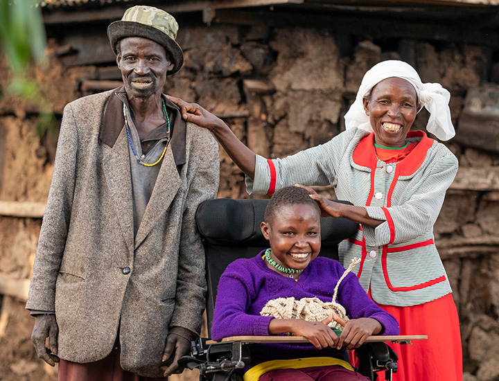 A man and a woman stand on either side of a girl sitting in a wheelchair, all smiling to the camera. The woman has one hand touching the girl's head, with her other hand on the man's shoulder.