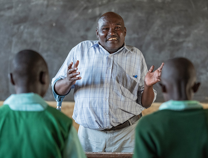 A man in a blue and white checkered shirt making a gesture with his hands while standing in front of a classroom, with one of his arms in an arm crutch.