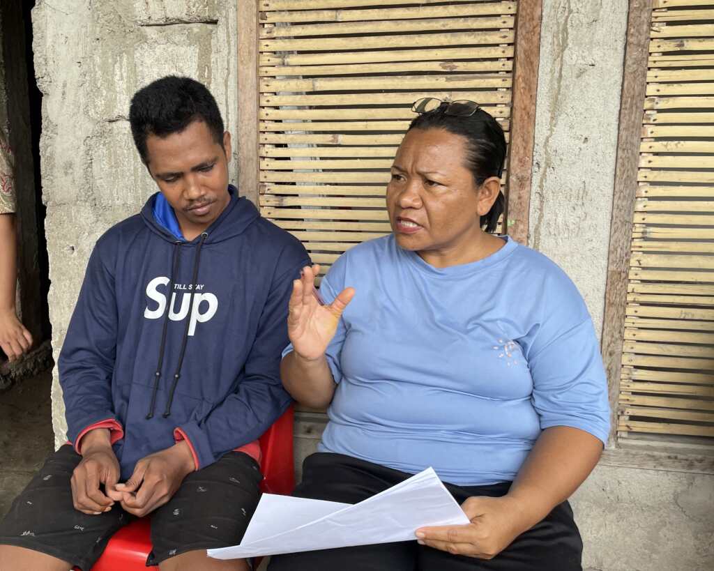Mama Novi is wearing a blue top and is seated beside a man wearing a dark blue hooded sweatshirt.