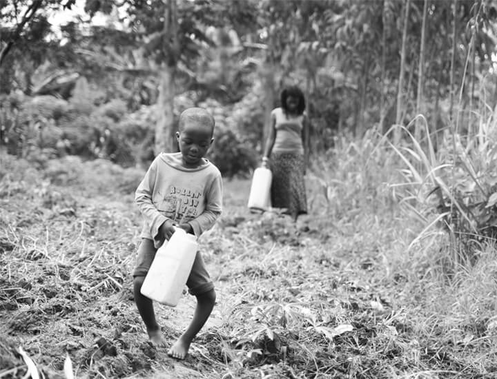 Child with a disability carries a heavy jug across rugged ground.