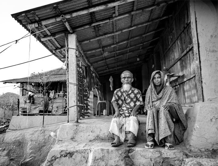 Older couple sit side by side outside in their village, both with concerned expressions.