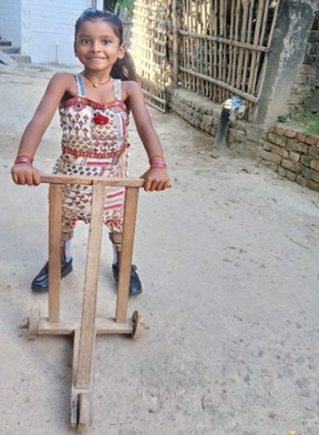 Sivani standing outside wearing her orthosis and holding onto a wooden mobility frame that has three wheels and helps her walk. She is smiling at the camera.