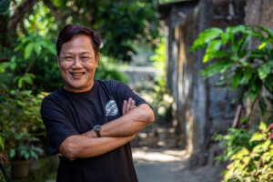 Sugeng, a participant in the CEPLERY project, is smiling with his arms folded. He is outside with green plants behind him.