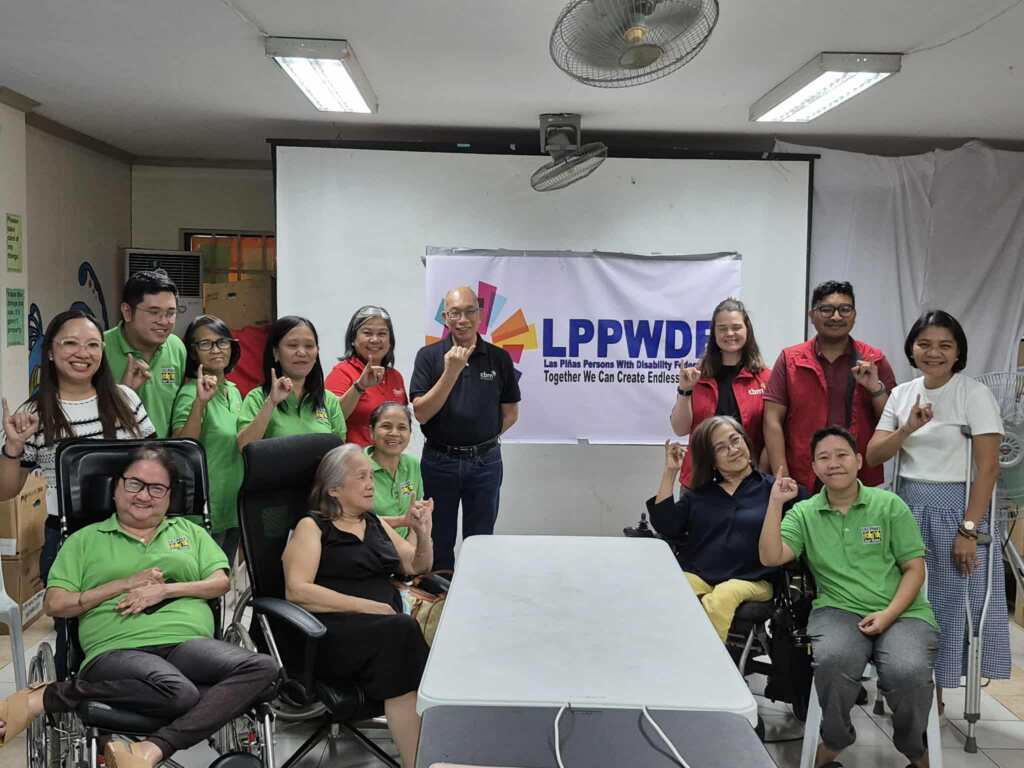 A group of people, including wheelchair users, pose together in a meeting room in front of a banner that reads “LPPWDF – Las Piñas Persons With Disability Federation, Inc. Together We Can Create Endless Possibilities.” Some wear green shirts, others red vests, and they are holding up one pinky finger.