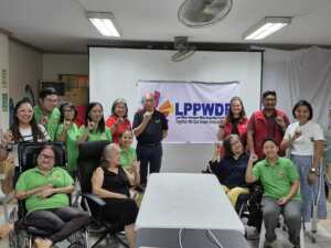 A group of people, including wheelchair users, pose together in a meeting room in front of a banner that reads “LPPWDF – Las Piñas Persons With Disability Federation, Inc. Together We Can Create Endless Possibilities.” Some wear green shirts, others red vests, and they are holding up one pinky finger.