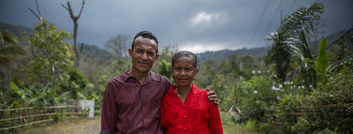 A man and women stand on a dirt road with green plants in the background. He has his arm around her and they are smiling.