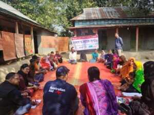 Members of the Village Savings and Loan Group sit outdoors. They are in a circle on an an orange mat.