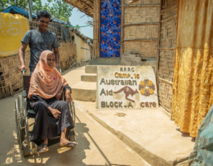 A women in a wheelchair stands next to a sign reading "Australian Aid, Block A"